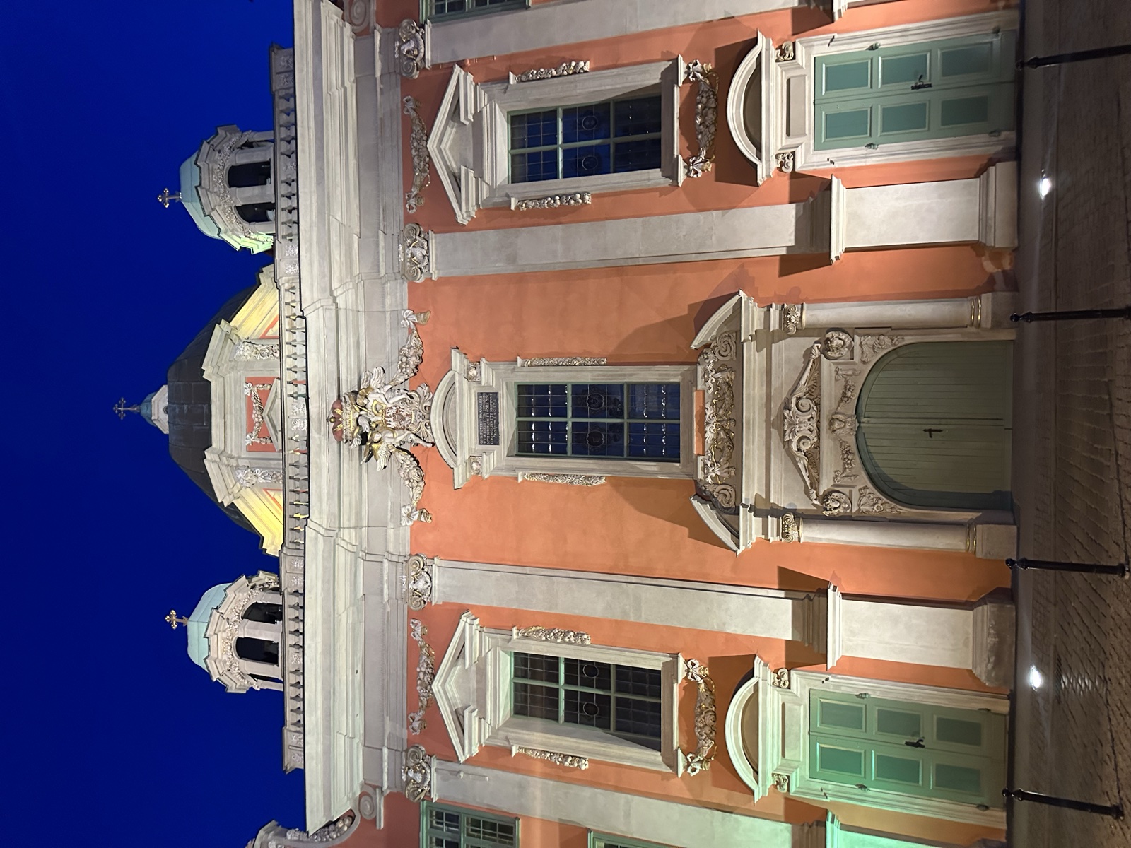 A baroque civic building at night, salmon walls, ornate white window frames, twin cupolas
