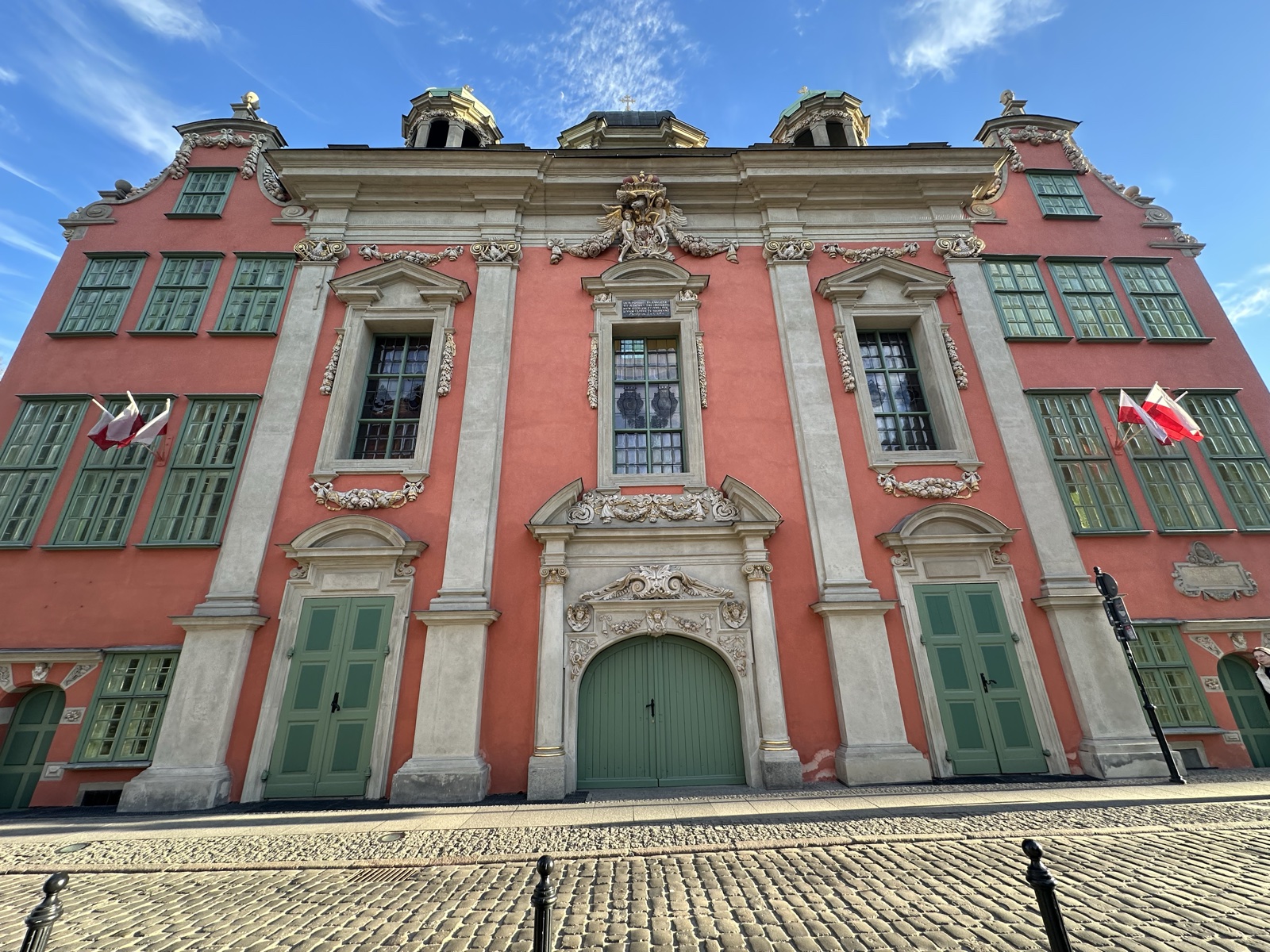 A salmon-pink baroque civic facade with white-framed windows, four cupolas, and Polish flags
